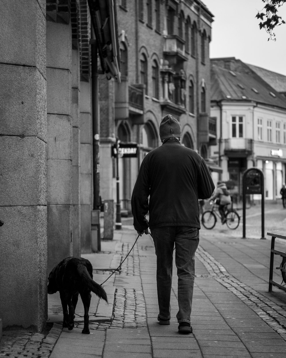 man in black jacket walking with black dog on sidewalk during daytime