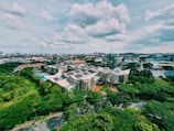 An aerial view of a complex of modern buildings with green roofs and solar panels, surrounded by lush greenery. The surrounding area features a mix of industrial and residential buildings, under a partly cloudy sky.