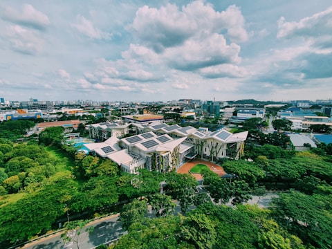 An aerial view of a complex of modern buildings with green roofs and solar panels, surrounded by lush greenery. The surrounding area features a mix of industrial and residential buildings, under a partly cloudy sky.
