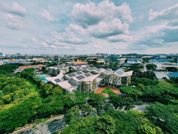 An aerial view of a complex of modern buildings with green roofs and solar panels, surrounded by lush greenery. The surrounding area features a mix of industrial and residential buildings, under a partly cloudy sky.