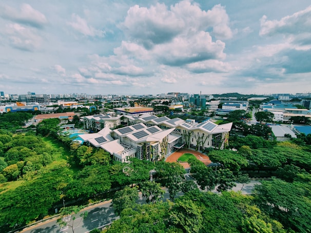 An aerial view of a complex of modern buildings with green roofs and solar panels, surrounded by lush greenery. The surrounding area features a mix of industrial and residential buildings, under a partly cloudy sky.