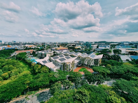 An aerial view of a complex of modern buildings with green roofs and solar panels, surrounded by lush greenery. The surrounding area features a mix of industrial and residential buildings, under a partly cloudy sky.