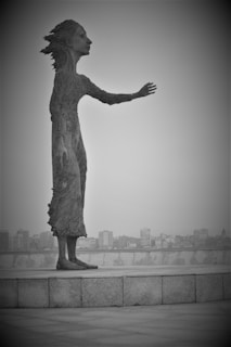 A large, textured sculpture of a person standing on a platform is prominently displayed. The figure appears to be reaching out with one arm extended. The background reveals a city skyline with numerous buildings visible in the distance. The image is in black and white, creating a stark, dramatic effect.