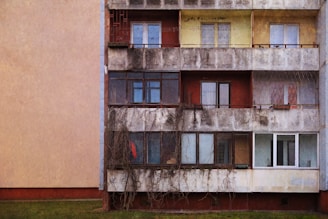 An old, weathered apartment building with multiple floors and balconies is shown. The facade appears worn and discolored due to age and exposure to the elements. Some balconies have glass enclosures, while others are open and feature metal railings. There are visible signs of neglect, including vines growing up the walls and some cracked or peeling paint. The windows vary in condition, with some appearing to have curtains or coverings, and others looking bare.