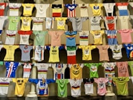 A collection of colorful cycling jerseys is displayed hanging on rows of lines. The jerseys feature various designs, logos, and brands, creating a vibrant and eclectic visual. Each jersey has distinct colors, patterns, and sponsorship logos, highlighting a broad spectrum of cycling history and culture.