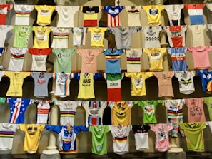 A collection of colorful cycling jerseys is displayed hanging on rows of lines. The jerseys feature various designs, logos, and brands, creating a vibrant and eclectic visual. Each jersey has distinct colors, patterns, and sponsorship logos, highlighting a broad spectrum of cycling history and culture.