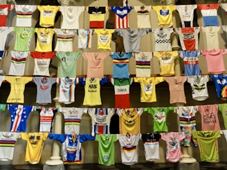 A collection of colorful cycling jerseys is displayed hanging on rows of lines. The jerseys feature various designs, logos, and brands, creating a vibrant and eclectic visual. Each jersey has distinct colors, patterns, and sponsorship logos, highlighting a broad spectrum of cycling history and culture.