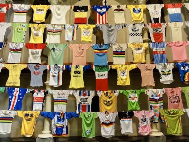 A collection of colorful cycling jerseys is displayed hanging on rows of lines. The jerseys feature various designs, logos, and brands, creating a vibrant and eclectic visual. Each jersey has distinct colors, patterns, and sponsorship logos, highlighting a broad spectrum of cycling history and culture.