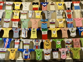 A collection of colorful cycling jerseys is displayed hanging on rows of lines. The jerseys feature various designs, logos, and brands, creating a vibrant and eclectic visual. Each jersey has distinct colors, patterns, and sponsorship logos, highlighting a broad spectrum of cycling history and culture.