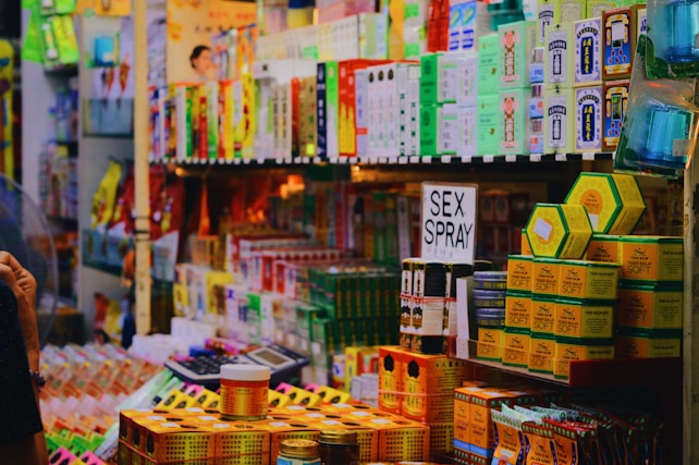 A cluttered store shelf displaying a variety of packaged products, mostly health and body care items. There's a prominent sign labeled 'SEX SPRAY' among the assortment of Tiger Balm products and other colorful boxes stacked neatly. The background is lined with more items, creating a busy and tightly packed retail environment.