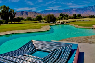 A serene landscape features a turquoise swimming pool bordered by a stone pathway and lush green grass. In the foreground, metallic geometric art sits on a platform, reflecting the light. The background features rolling hills and a mountain range under a bright blue sky with wispy clouds.