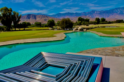 A serene landscape features a turquoise swimming pool bordered by a stone pathway and lush green grass. In the foreground, metallic geometric art sits on a platform, reflecting the light. The background features rolling hills and a mountain range under a bright blue sky with wispy clouds.