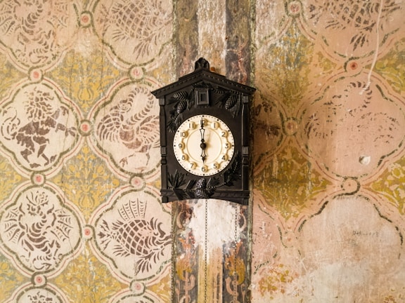An ornate black wooden wall clock hangs against a vintage wallpaper backdrop featuring a repetitive floral pattern in muted tones of beige, yellow, and green. The clock displays Roman numerals and has decorative carvings on its body.