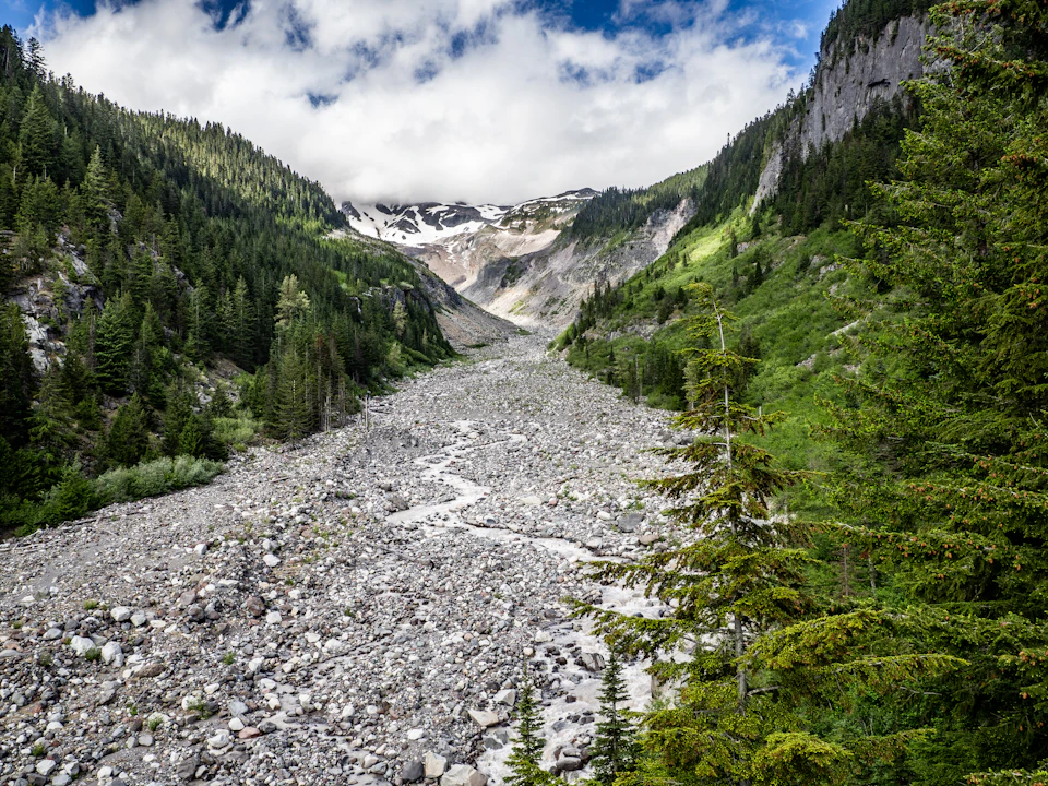 The image depicts a breathtaking view of a mountain range with a river flowing through it, nestled amidst lush green trees and shrubs. The mountains are covered in snow, indicating that they are located at a high altitude. The sky above is clear blue with fluffy white clouds scattered across the expanse. The perspective of the photo is from a higher vantage point, providing an expansive view of the natural landscape below.