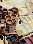 A series of circular and rectangular stone pits filled with liquids of various colors, likely used for dyeing fabrics. The stones are weathered and earth-toned while the liquids are in shades of yellow, blue, and pink. Fabrics and ropes are laid over some of the pits, and there is a solitary garment soaking. The arrangement suggests a traditional dyeing or tanning process.