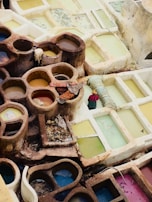 A series of circular and rectangular stone pits filled with liquids of various colors, likely used for dyeing fabrics. The stones are weathered and earth-toned while the liquids are in shades of yellow, blue, and pink. Fabrics and ropes are laid over some of the pits, and there is a solitary garment soaking. The arrangement suggests a traditional dyeing or tanning process.