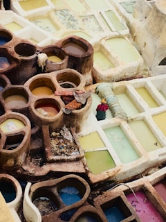 A series of circular and rectangular stone pits filled with liquids of various colors, likely used for dyeing fabrics. The stones are weathered and earth-toned while the liquids are in shades of yellow, blue, and pink. Fabrics and ropes are laid over some of the pits, and there is a solitary garment soaking. The arrangement suggests a traditional dyeing or tanning process.