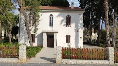 A two-story white house with brown shutters and a red tile roof is surrounded by trees and greenery. The entrance features a wooden door with a small overhang, and the property is enclosed by a low stone fence with a wooden gate. The setting is a well-maintained garden area with some palm trees and other vegetation.