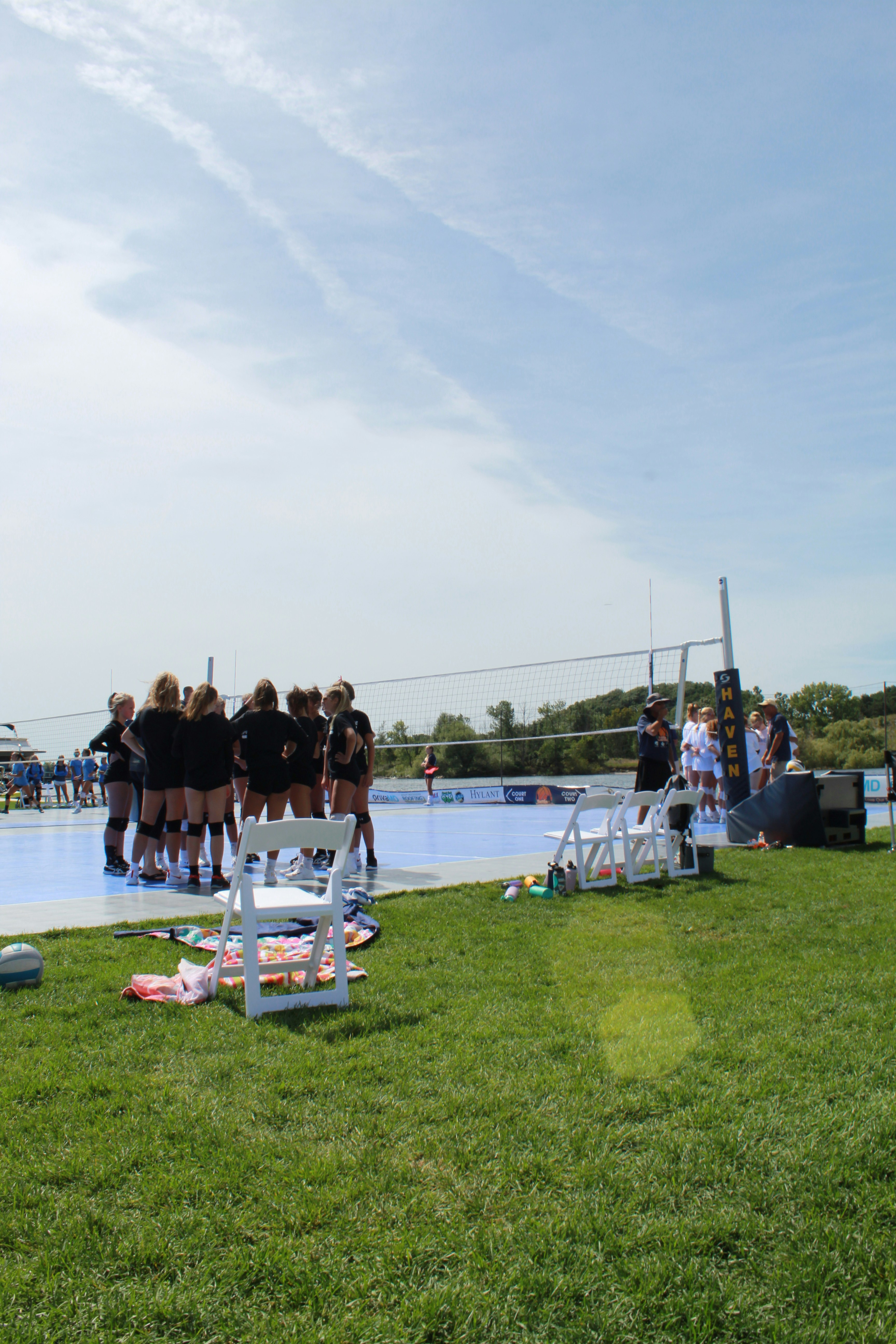 A group of people wearing sports uniforms are gathered in a huddle on a volleyball court. The court is set outdoors on a grassy field under a mostly clear sky. Various items such as water bottles and personal belongings are scattered on the grass, with some white chairs placed nearby.