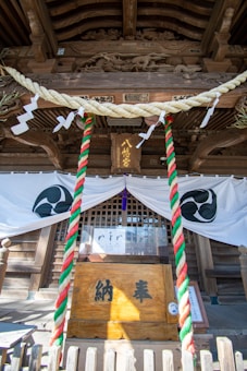 An ornate wooden structure features intricate carvings, including a dragon motif, under a traditional Japanese temple roof. A large offering box with Japanese characters sits in front, flanked by colorful twisted ropes in red, white, and green. Decorative white flags and a purple tassel hang above, while a decorative banner with circular symbols drapes across the entrance.