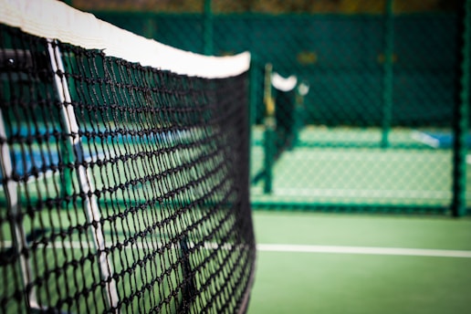 A close-up of a tennis ball resting on a vibrant green tennis court, with the net softly blurred in the background.