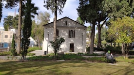 A quaint white building surrounded by tall trees and greenery, with a well-maintained garden in the foreground. A person sits on a park bench, appearing relaxed and engaged with a book or device.