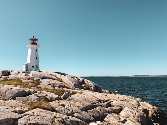 white and red lighthouse on brown rocky shore under blue sky during daytime in Peggy's Point Lighthouse Canada