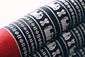 The image features a fabric with intricate patterns in white woven onto a black background. The patterns include geometric shapes and representations of animals or objects, repeated in horizontal rows. The lower portion of the fabric is a solid red color, providing contrast to the patterned section.