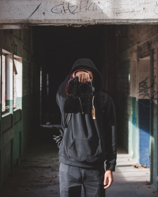 A person wearing a dark hoodie is standing in a dimly lit, abandoned corridor. They are holding a camera up to their face, pointing it towards the viewer. The walls are covered in graffiti, and sunlight filters through partially boarded windows.