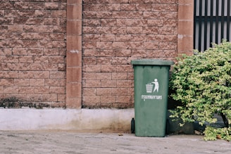 A large green trash bin with white text and a symbol stands beside a leafy bush. The background features a textured brick wall with a vertical column dividing sections of the wall.