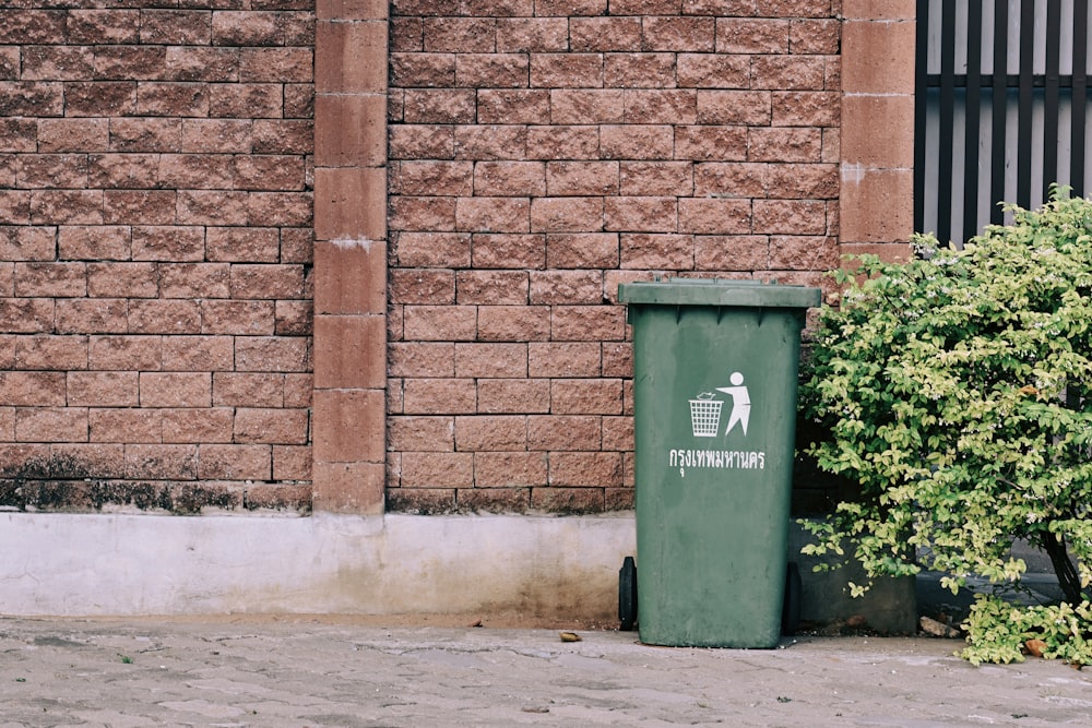 Cubo de basura verde junto a la pared de ladrillo marrón