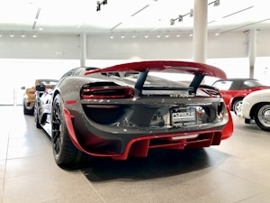 A sleek, high-performance sports car is prominently displayed indoors, highlighting its glossy finish and aerodynamic design. Surrounding it are other luxurious cars, suggesting a showroom setting.