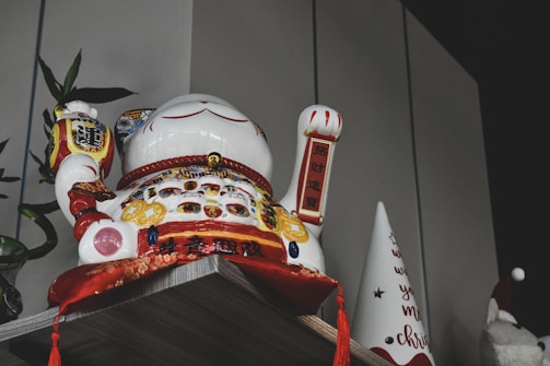 A decorative ceramic cat figurine is positioned on a shelf, featuring a raised paw and colorful designs with gold and red accents. Surrounding the figurine is a small plant with green leaves and a white cone-shaped object with red text.