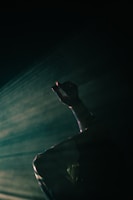 A dimly lit scene highlights a raised hand with dramatic shadows and streams of light suggesting movement. The atmosphere is moody and introspective.