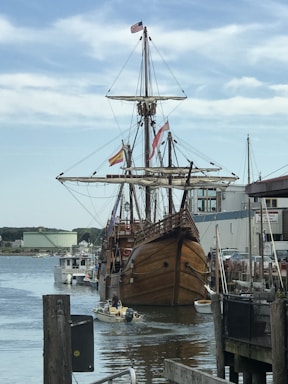 A large wooden sailing ship with multiple sails is docked at a marina. There are various flags, including an American flag, flying from its masts. Several smaller boats and a waterfront with some buildings and docks are visible in the background. The sky is partly cloudy.