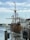 A large wooden sailing ship with multiple sails is docked at a marina. There are various flags, including an American flag, flying from its masts. Several smaller boats and a waterfront with some buildings and docks are visible in the background. The sky is partly cloudy.