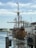 A large wooden sailing ship with multiple sails is docked at a marina. There are various flags, including an American flag, flying from its masts. Several smaller boats and a waterfront with some buildings and docks are visible in the background. The sky is partly cloudy.