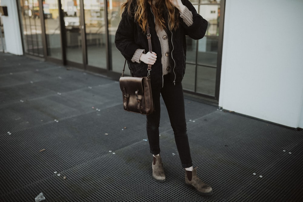 woman in black leather jacket and black pants holding brown leather sling bag