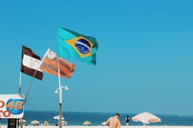 A beach scene with three flags flying prominently against a clear blue sky. The flags include the Brazilian national flag and two others featuring distinct designs. In the background, beachgoers are visible near colorful parasols, enjoying a bright day by the sea.