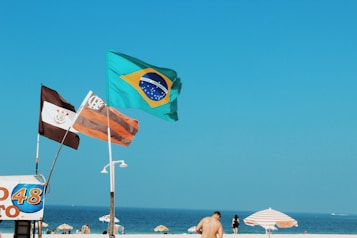 A beach scene with three flags flying prominently against a clear blue sky. The flags include the Brazilian national flag and two others featuring distinct designs. In the background, beachgoers are visible near colorful parasols, enjoying a bright day by the sea.