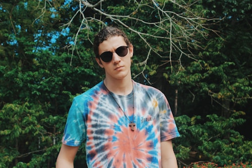 A person wearing sunglasses and a colorful tie-dye t-shirt stands in front of lush green foliage. They have short, curly hair and are wearing a necklace with a pendant. The background consists of dense trees with branches and leaves.