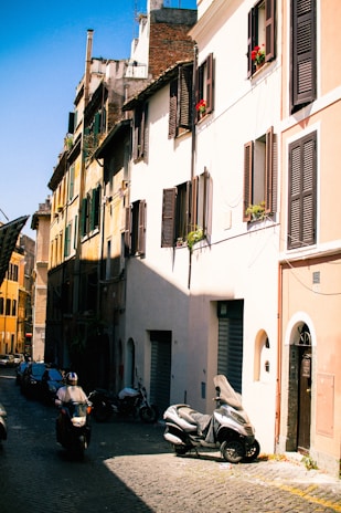 A lively street in Rome with cobblestones, scooters, and historic buildings bathed in warm sunlight.