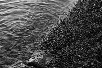 A black and white photograph of a rocky shoreline with small waves gently lapping against the pebbles.