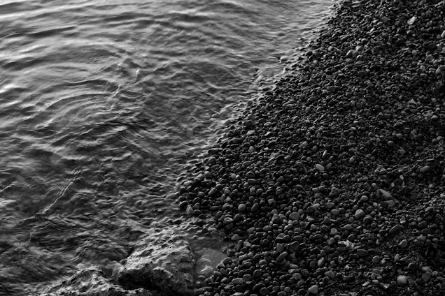 A black and white photograph of a rocky shoreline with small waves gently lapping against the pebbles.