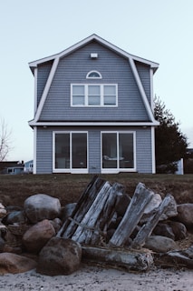 A two-story house with gray shingle siding and large windows is situated on a grassy area. In the foreground, there are weathered wooden logs and large rocks, possibly part of a natural or constructed barrier along a beach.