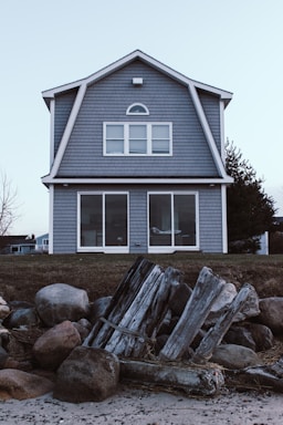 A two-story house with gray shingle siding and large windows is situated on a grassy area. In the foreground, there are weathered wooden logs and large rocks, possibly part of a natural or constructed barrier along a beach.