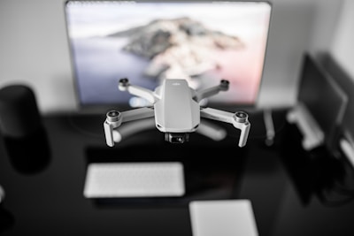 A small drone hovers in front of a computer screen in an office setting. The background includes a blurred desktop with a keyboard, a tablet, and other electronic devices.