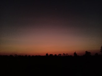 A low-light scene depicting a sunset or sunrise with a clear gradient from deep orange near the horizon, transitioning to darker shades of blue and black as it moves upward. Silhouetted trees and landscape elements appear in the lower portion of the image.