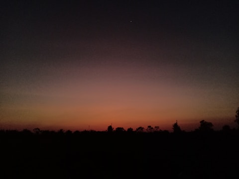 A low-light scene depicting a sunset or sunrise with a clear gradient from deep orange near the horizon, transitioning to darker shades of blue and black as it moves upward. Silhouetted trees and landscape elements appear in the lower portion of the image.