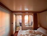 A cozy room with a neatly made bed and a window offering a view of a vibrant sunset over the ocean. Soft lighting creates a warm and inviting atmosphere, contrasting with the deep blue of the ocean. The room is simple, featuring neutral-colored walls and a curtain that partially covers the window, enhancing the intimate setting.